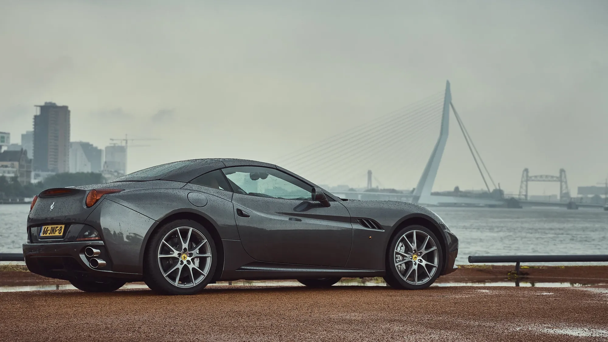 Grey sports car on the quay along the Maas in Rotterdam, with the Erasmus bridge behind it