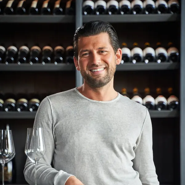 Man in a light sweater in front of a wine rack, wine glasses on the counter